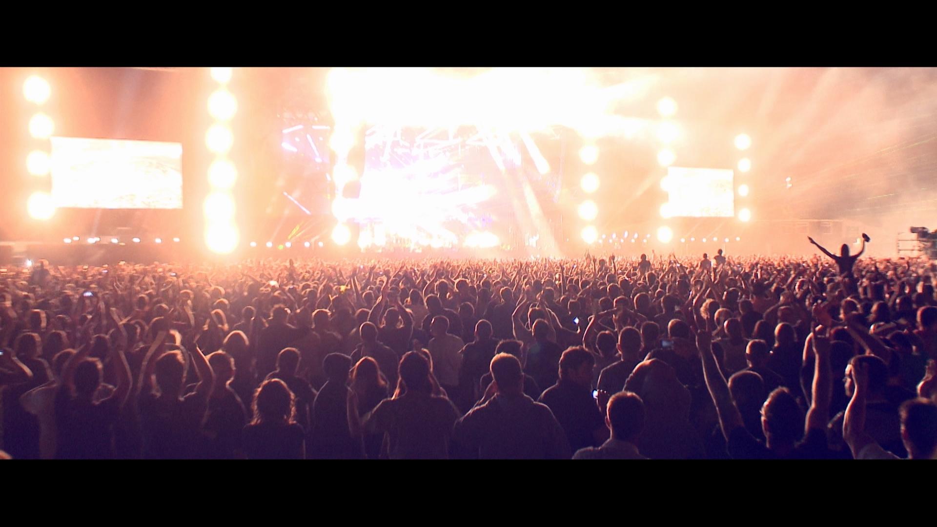 A large crowd of people at an outdoor concert with bright stage lighting and large screens in the background at night.