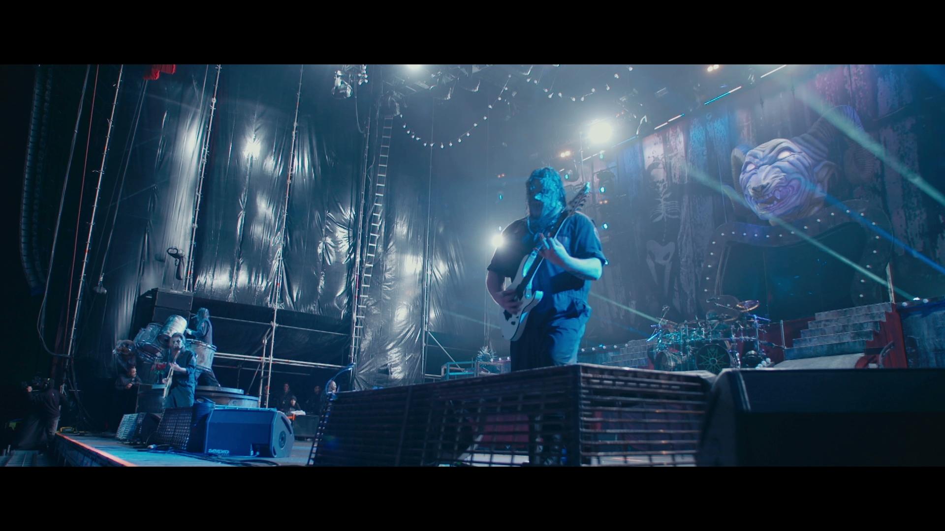 Slipknot guitarist performs on a dimly lit stage with blue lighting, surrounded by various musical equipment and decorations. The background features a large banner with artistic visuals.