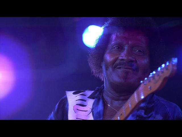 Albert Collins with an electric guitar smiles, illuminated by purple and blue stage lights.