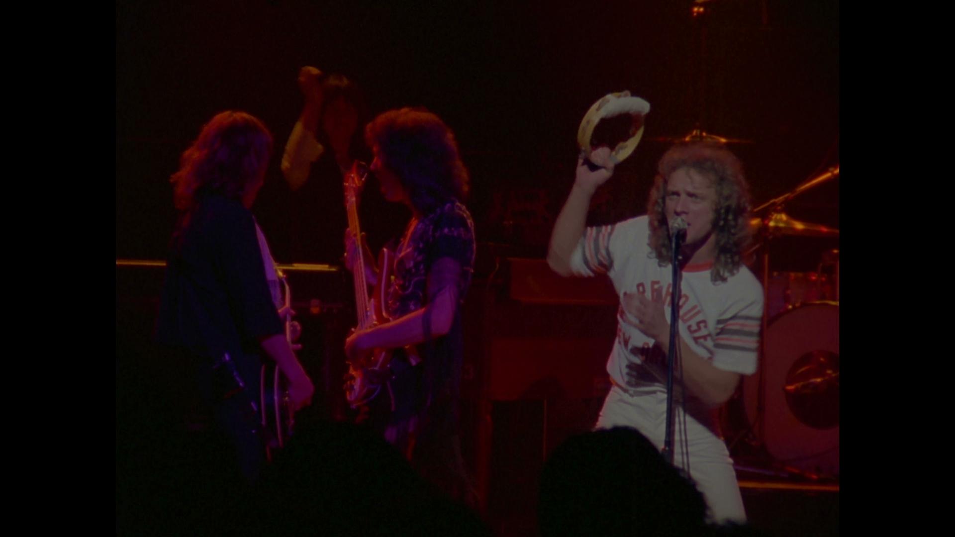 Foreigner singer holds a tambourine while performing on stage with a band. Two guitarists are in the background. The stage is dimly lit with red and purple lighting.