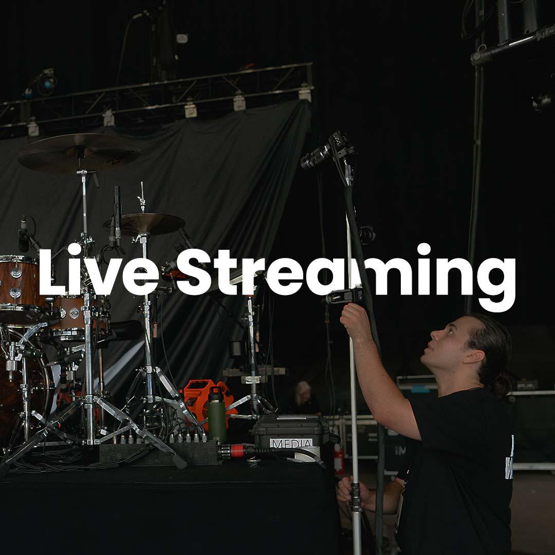 Person adjusting a microphone stand on stage with drum set in the background. Text overlay reads "Live Streaming.