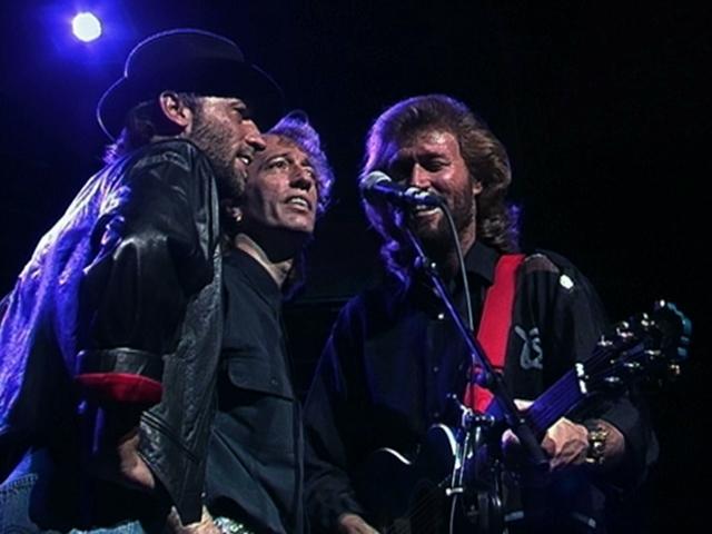 The Bee Gees are standing close together, singing into a microphone on stage under a spotlight. One of them is playing a guitar.