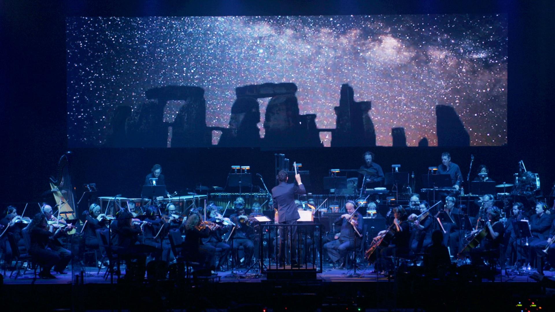 A symphony orchestra performs on stage in front of a large screen displaying a starry sky and Stonehenge, with musicians playing various instruments under blue lighting.