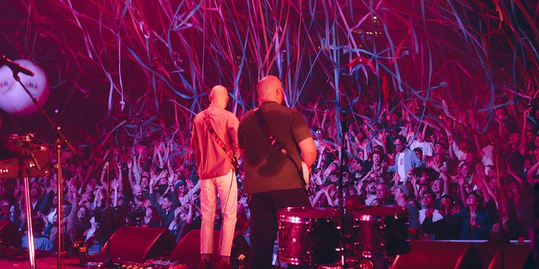 A Bombay Bicycle Club concert stage with two musicians playing guitars facing a large, enthusiastic crowd. Pink lighting and streamers fill the air.