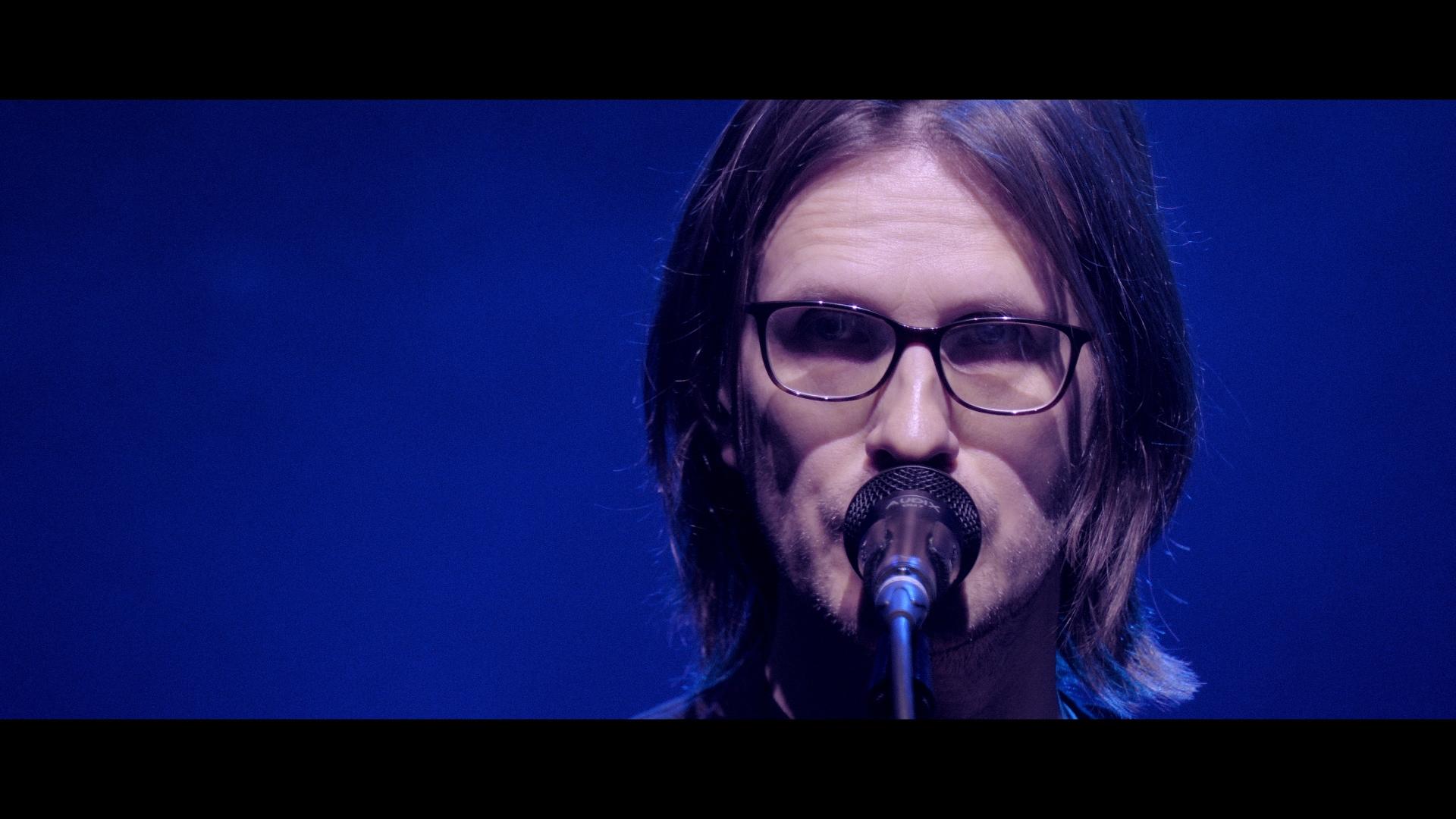 Steven Wilson with shoulder-length hair and glasses is speaking or singing into a microphone with a blue-lit background.