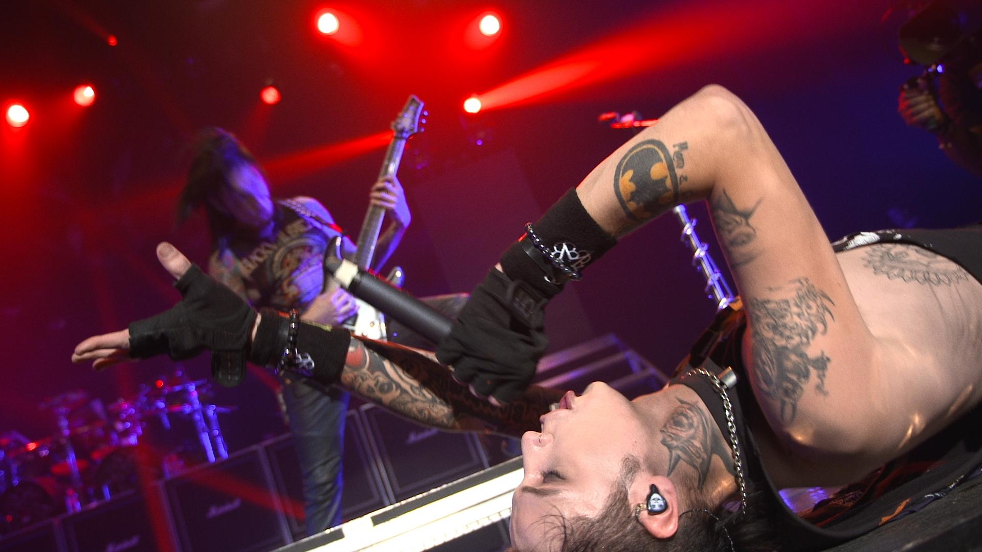 Black Veil Brides lying on stage singing into a microphone, another musician playing guitar in the background, red stage lights.