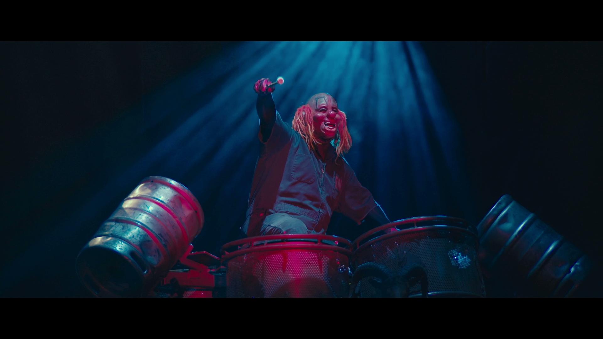 Slipknot clown with long hair energetically plays drums on stage under dramatic blue lighting.