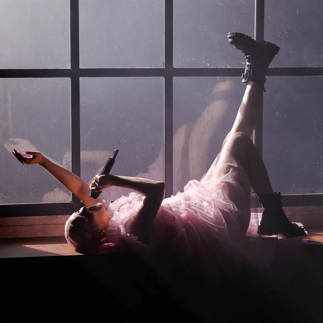 Anne-Marie in a pink dress and black boots, lying on their back with legs up against a window, holding a microphone, and bathed in soft light.