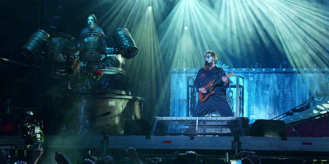 Slipknot performing on stage with intense lighting; one member plays the guitar while another is on an elevated platform with large drums. Both wear costumes and masks.