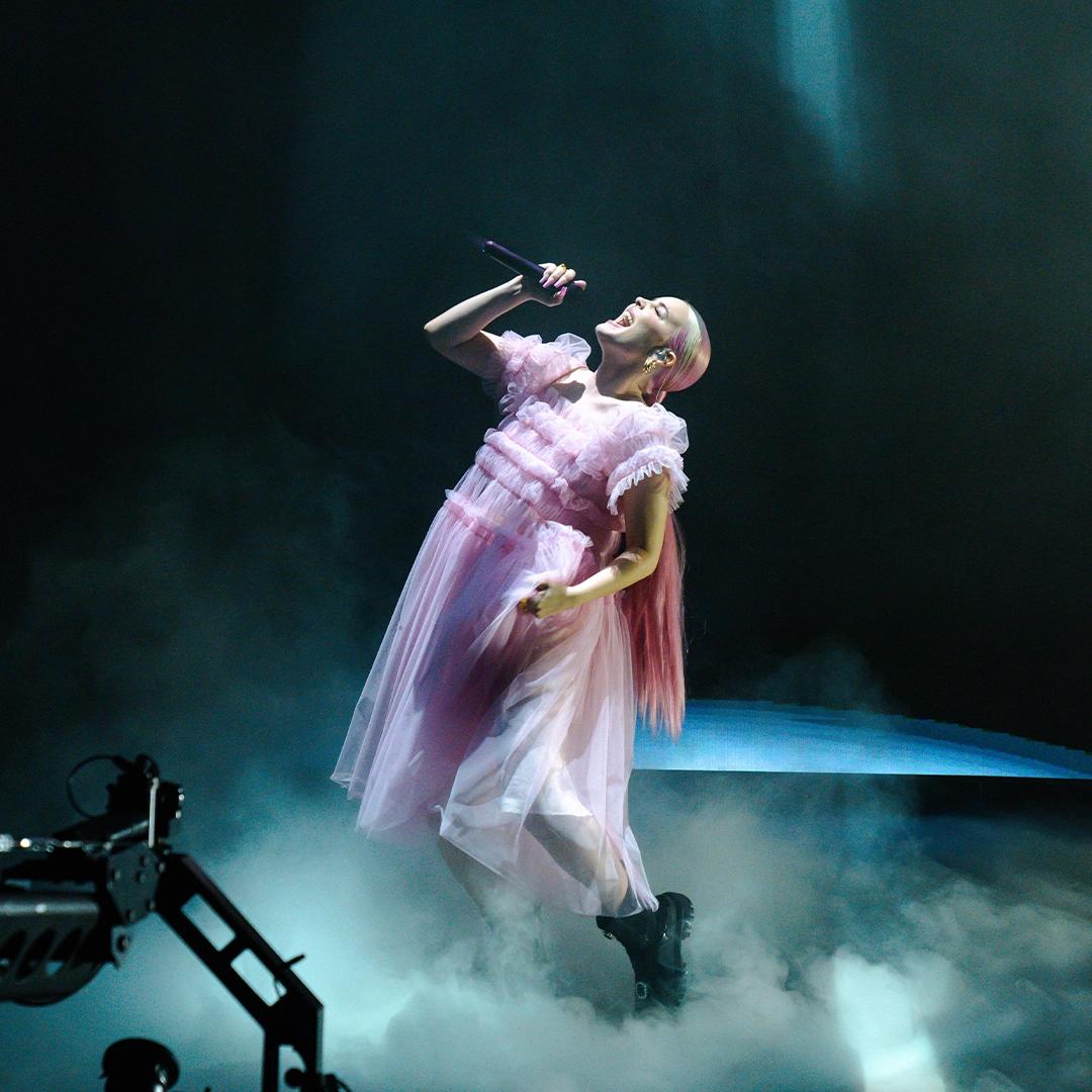 Anne-Marie in a pink dress sings passionately on stage with a microphone, surrounded by dramatic lighting and mist.