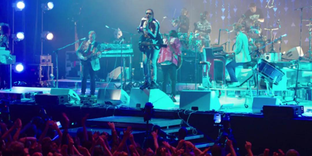 A band performs on stage with various instruments under blue lighting, as a crowd of fans with raised hands watches them from the front.