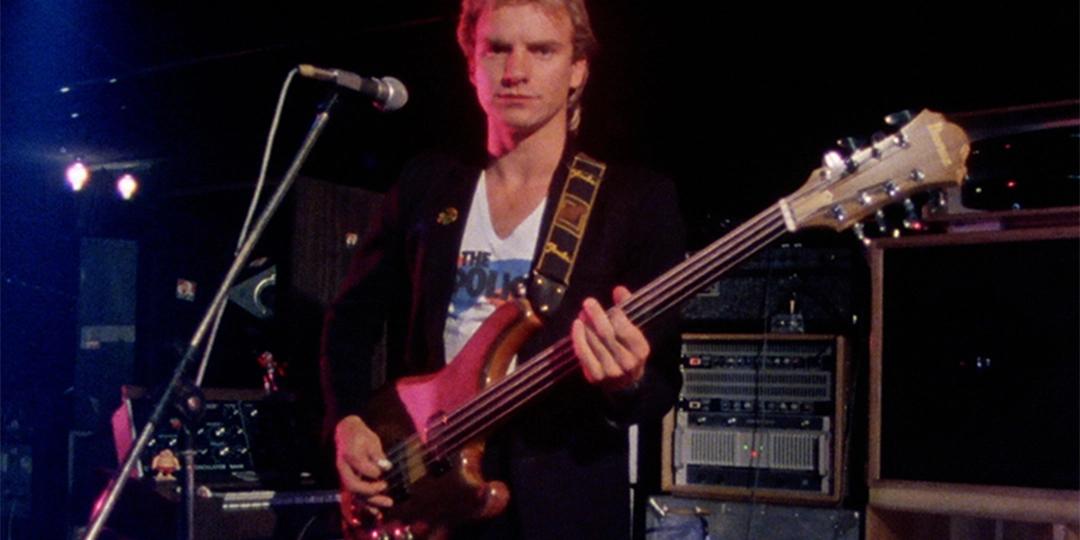 The Police bassist playing a bass guitar on stage, wearing a black jacket over a white T-shirt, standing in front of a microphone with a serious expression, with equipment and other instruments in the background.