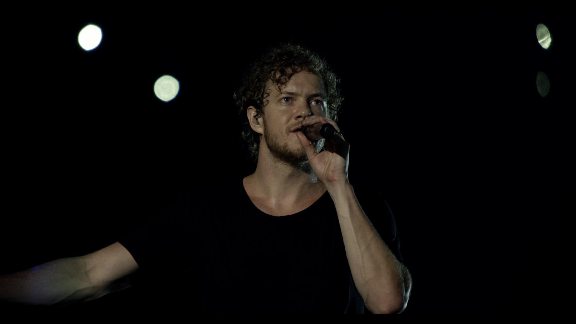 Imagine Dragons singer with curly hair and a beard sings into a microphone on a dark stage with bright lights in the background.