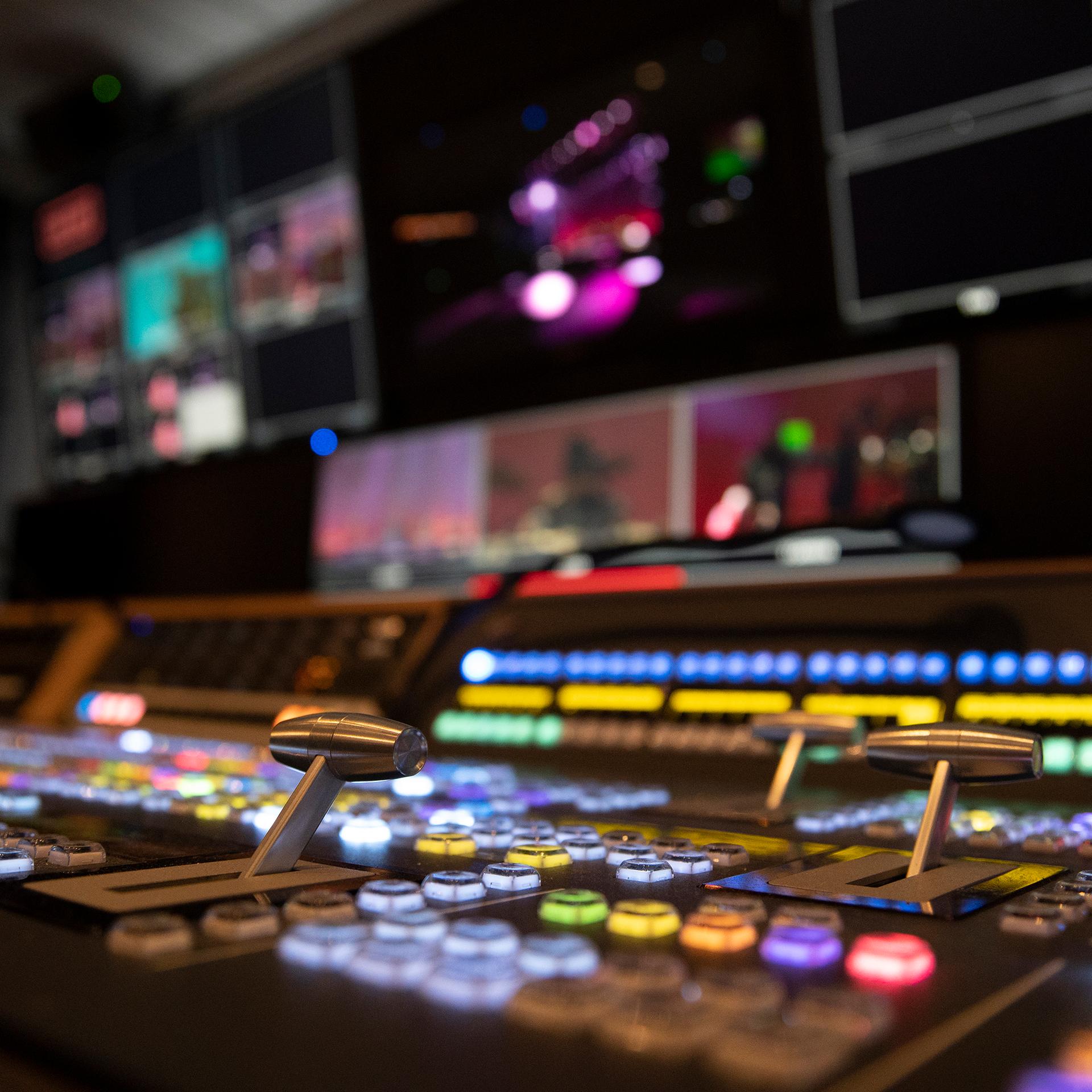 Close-up of a control panel with illuminated buttons and levers in a broadcast studio, multiple screens displaying video feeds in the background.