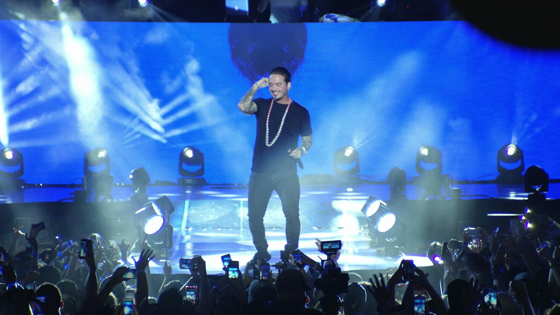 J Balvin on stage under blue, backlit lighting, with a crowd of people taking photos and videos with their phones.