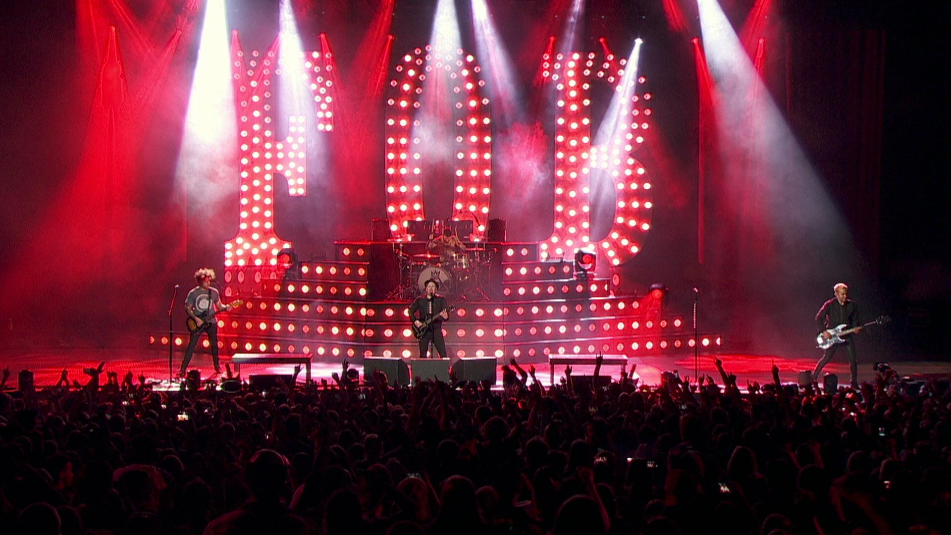 Fall Out Boy performs on stage with bright red and white lights, a large illuminated "FOB" sign, and a cheering crowd in the foreground.