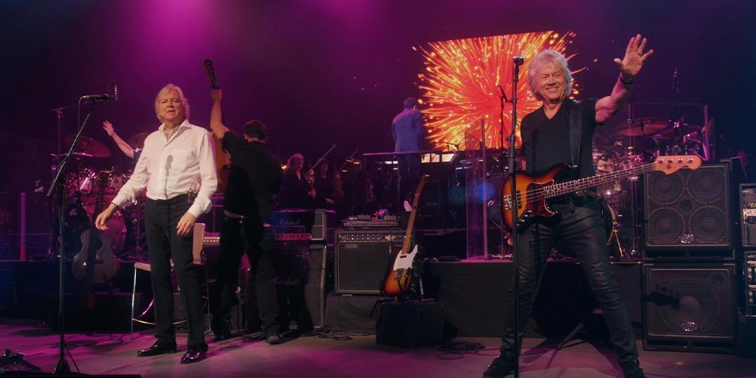 The Moody Blues perform on stage with vibrant pink and purple lights. The bassist waves to the audience, while another member stands with a microphone. A screen displays a burst of light in the background.