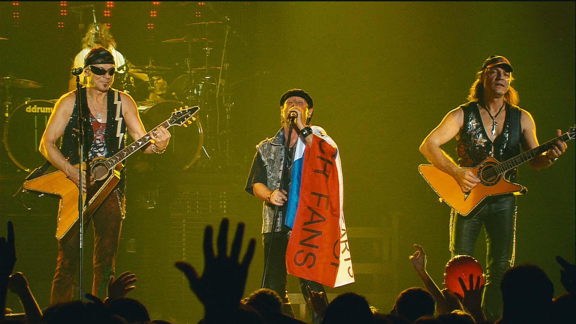 Scorpions perform on stage; the singer in the center holds a flag, while the other two play guitars. Fans' hands are visible in the foreground.