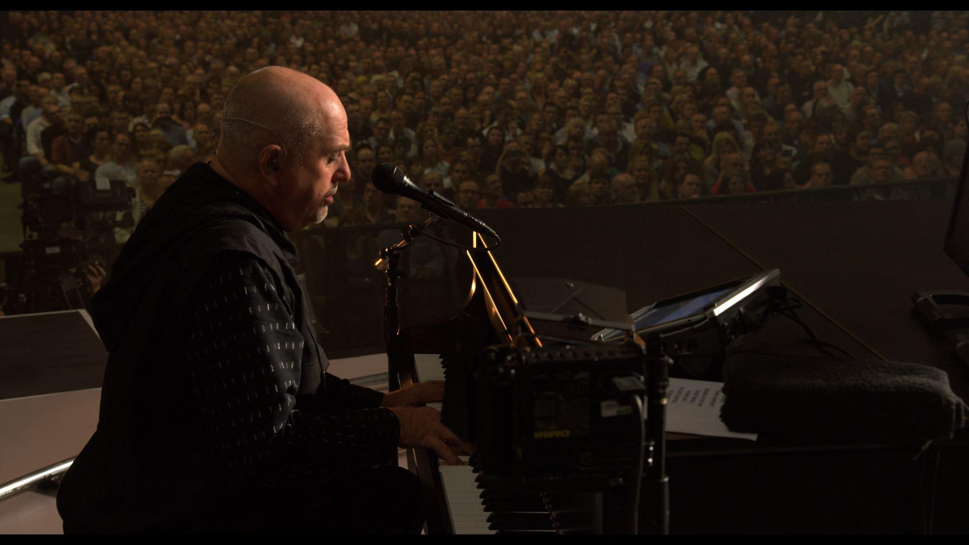 Peter Gabriel plays the piano and sings into a microphone facing an audience in a packed venue.