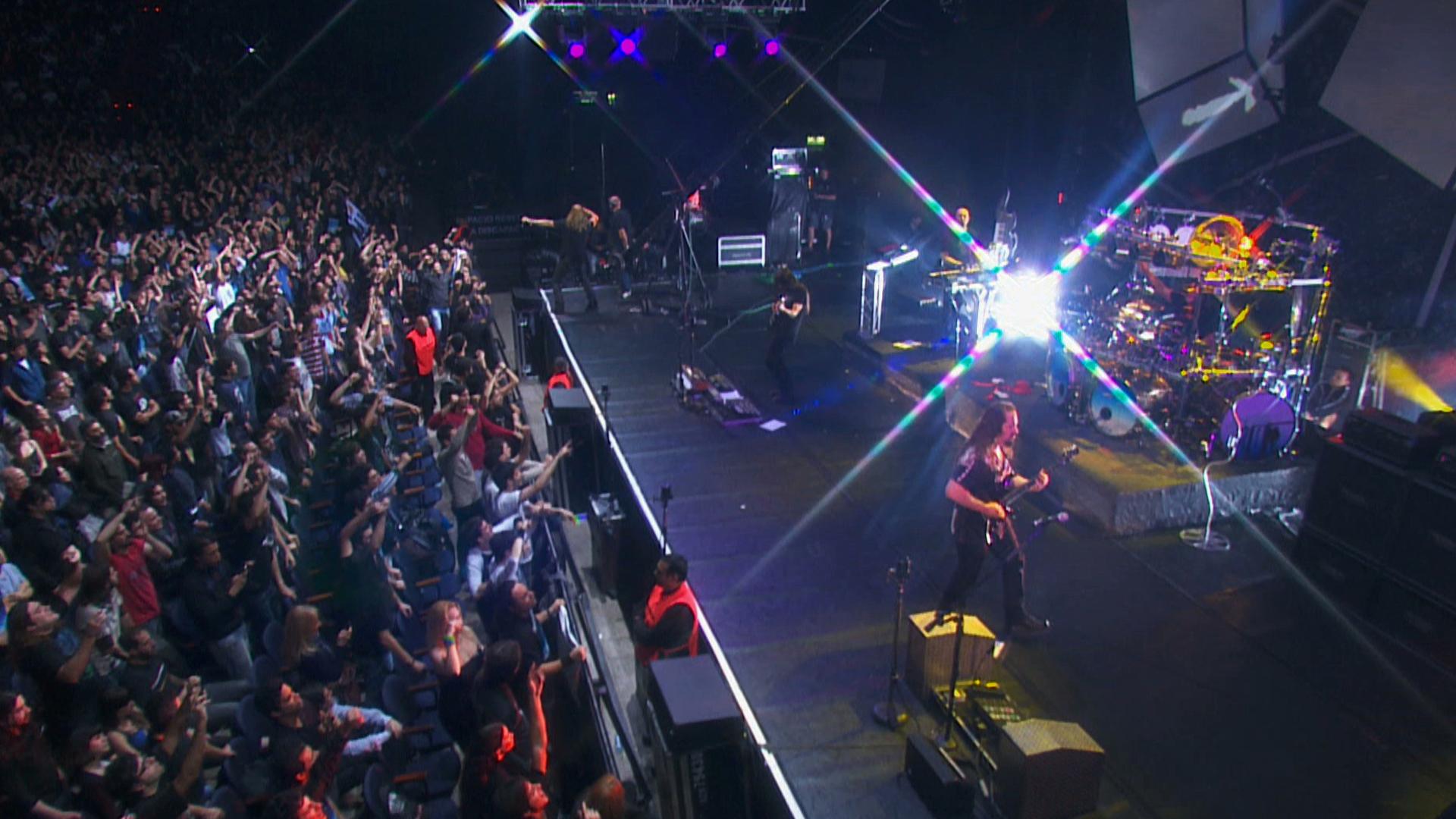 dream theater performs live on stage with a large audience visible on the left side of the image, cheering and clapping. Musical equipment and bright stage lights are also present.