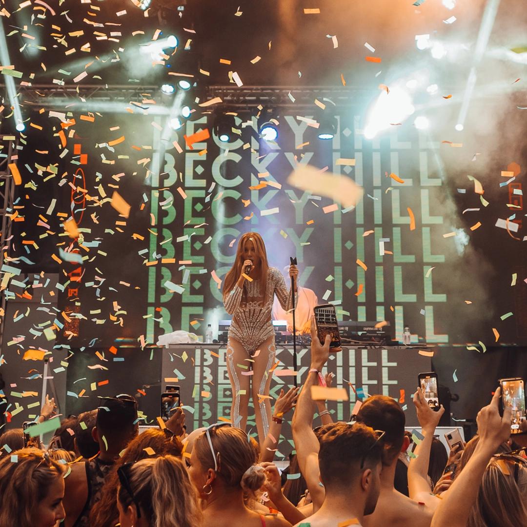 Becky Hill performs on stage surrounded by cheering audience and confetti, with the name "Becky Hill" displayed in the background.