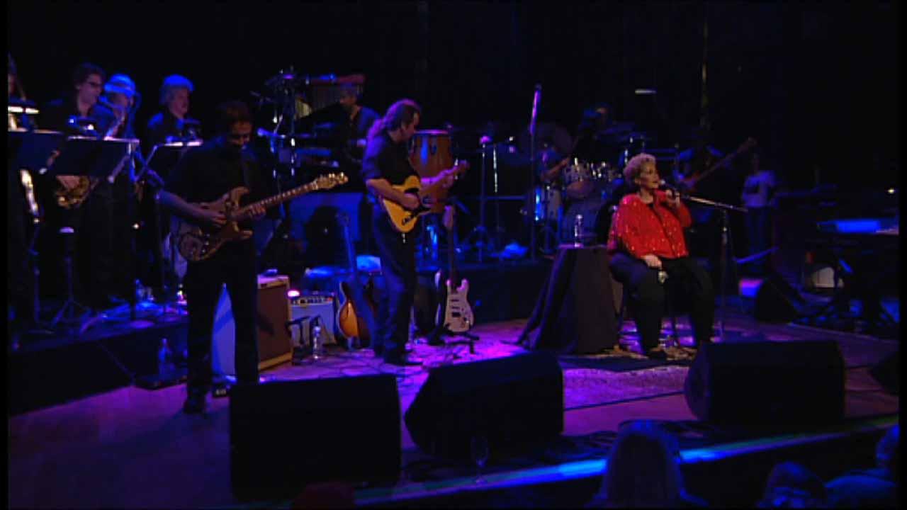 Etta James performs on stage with multiple musicians playing guitars, drums, and brass instruments. A person in a red shirt sits and sings into a microphone. The stage is dimly lit.