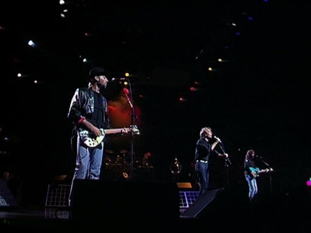 Bee Gees perform on a dimly lit stage. Three members are visible, each playing a guitar and standing at microphones. Colorful lights illuminate the background.