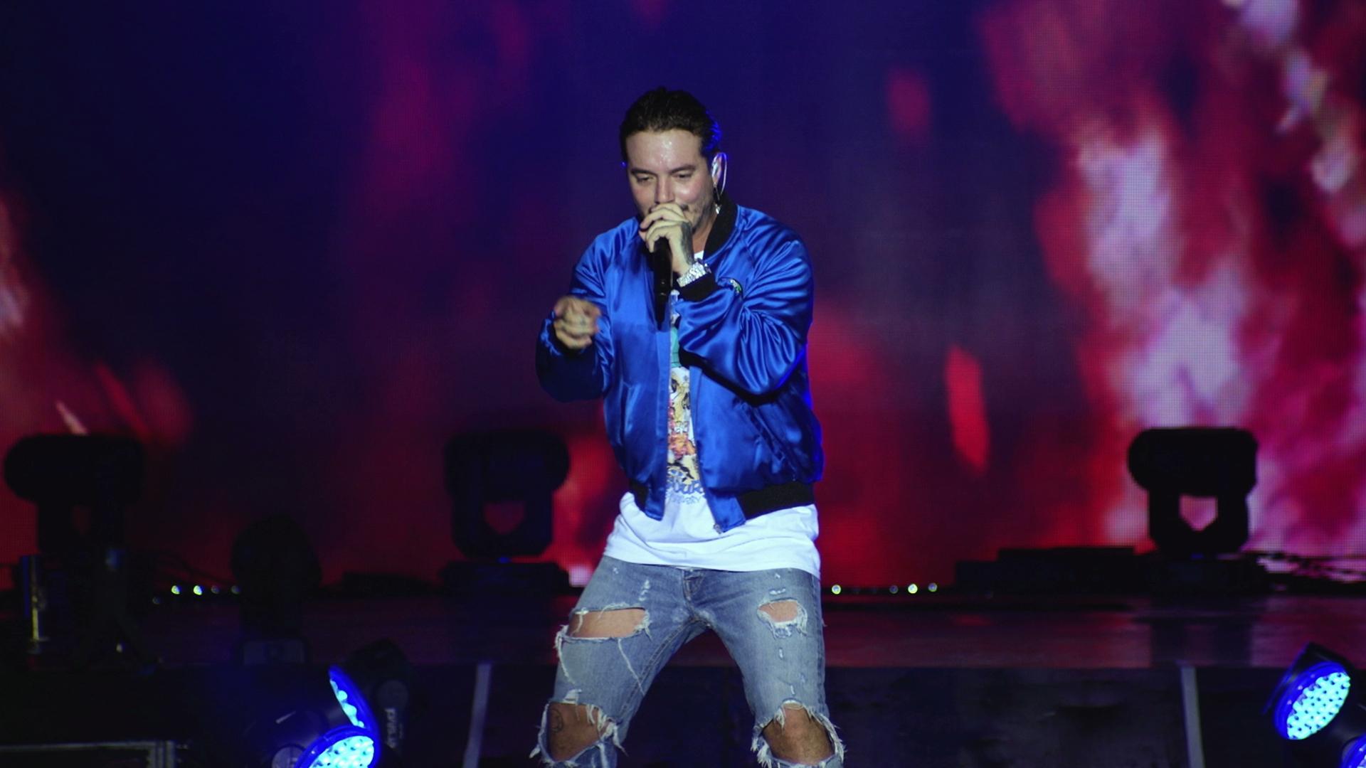 J Balvin wearing a blue jacket and ripped jeans performs on stage with a microphone, against a dark, colorful background.