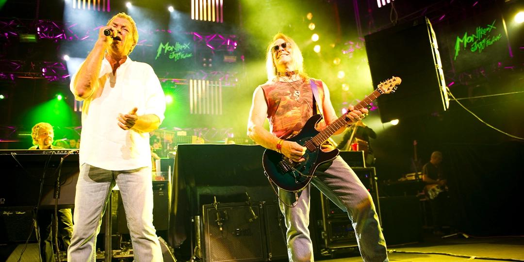 Deep Purple perform on stage under vibrant lighting, one singing into a microphone while the other plays an electric guitar. In the background, a keyboardist is partially visible.