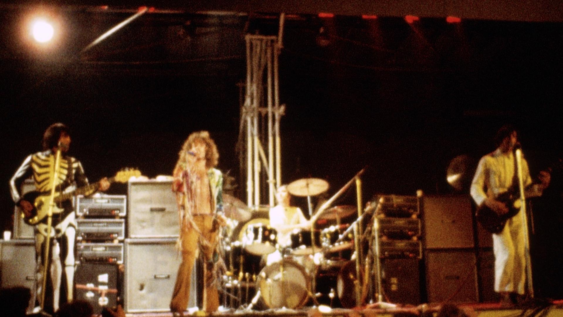 The Who perform on stage with their instruments, one holding a bass guitar, another singing into a microphone, and the third playing drums, under stage lighting.