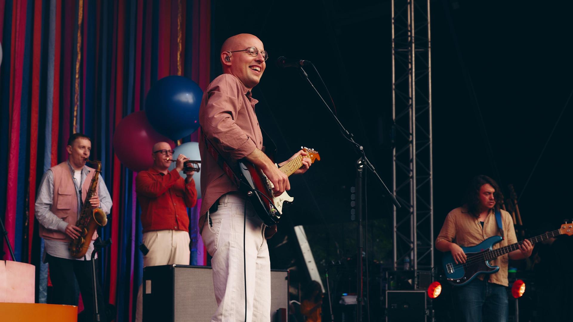 Bombay Bicycle Club frontman Jack in a pink shirt plays an electric guitar and sings into a microphone on stage. A saxophonist, trumpeter, and bassist perform with him, with colorful balloons in the background.