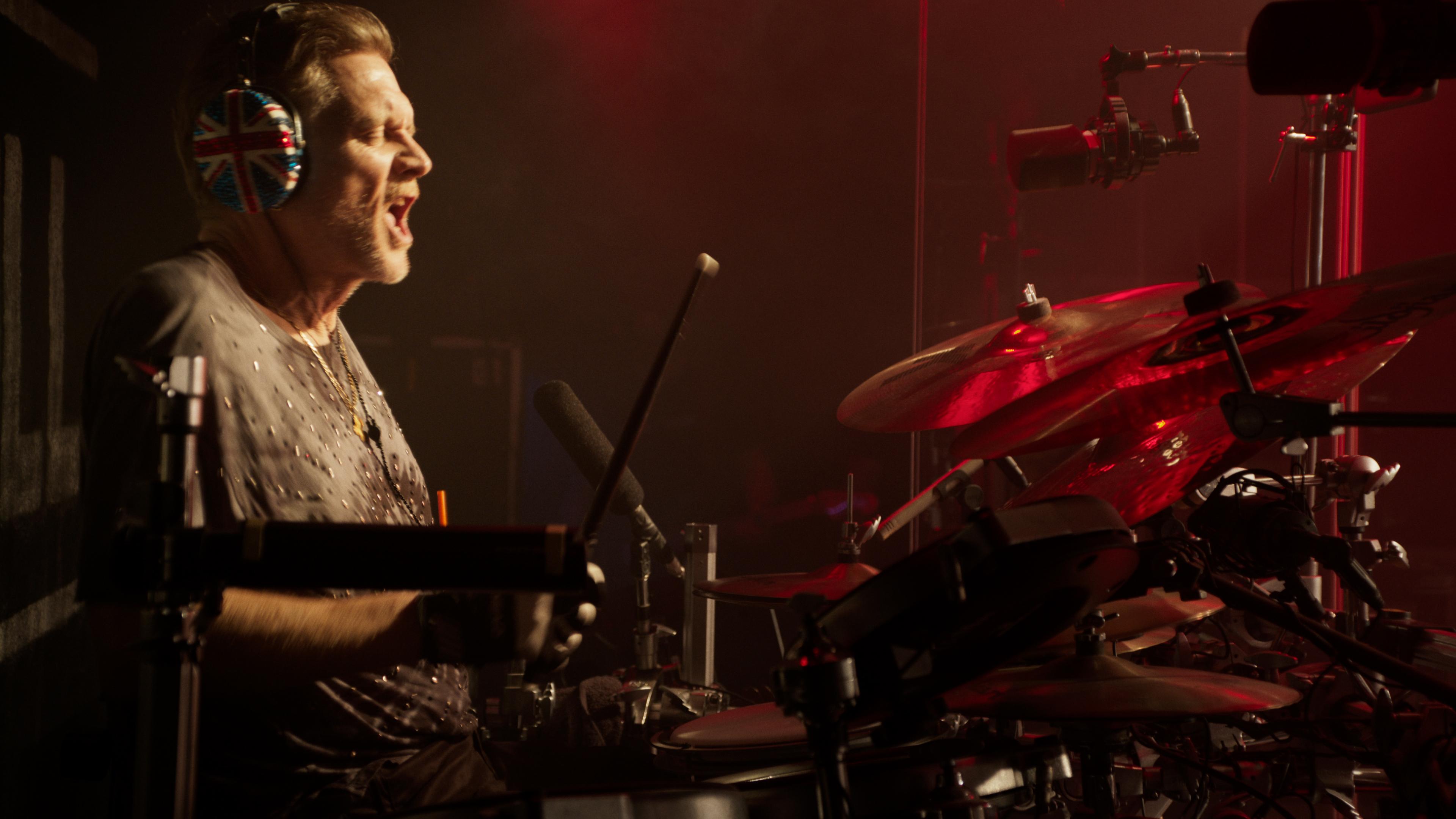 Def Leppard drummer wearing headphones with a Union Jack design passionately plays a drum set on stage, illuminated by red lighting.