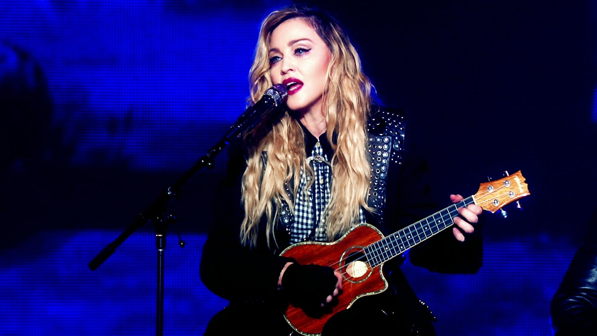 Madonna with long blonde hair performs at concert on stage, singing into a microphone while playing a small guitar.