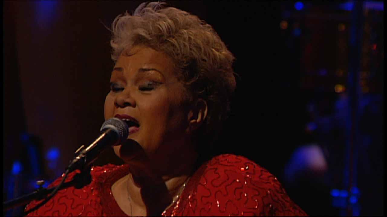 Etta James with short, curly hair sings into a microphone while wearing a red outfit on stage.