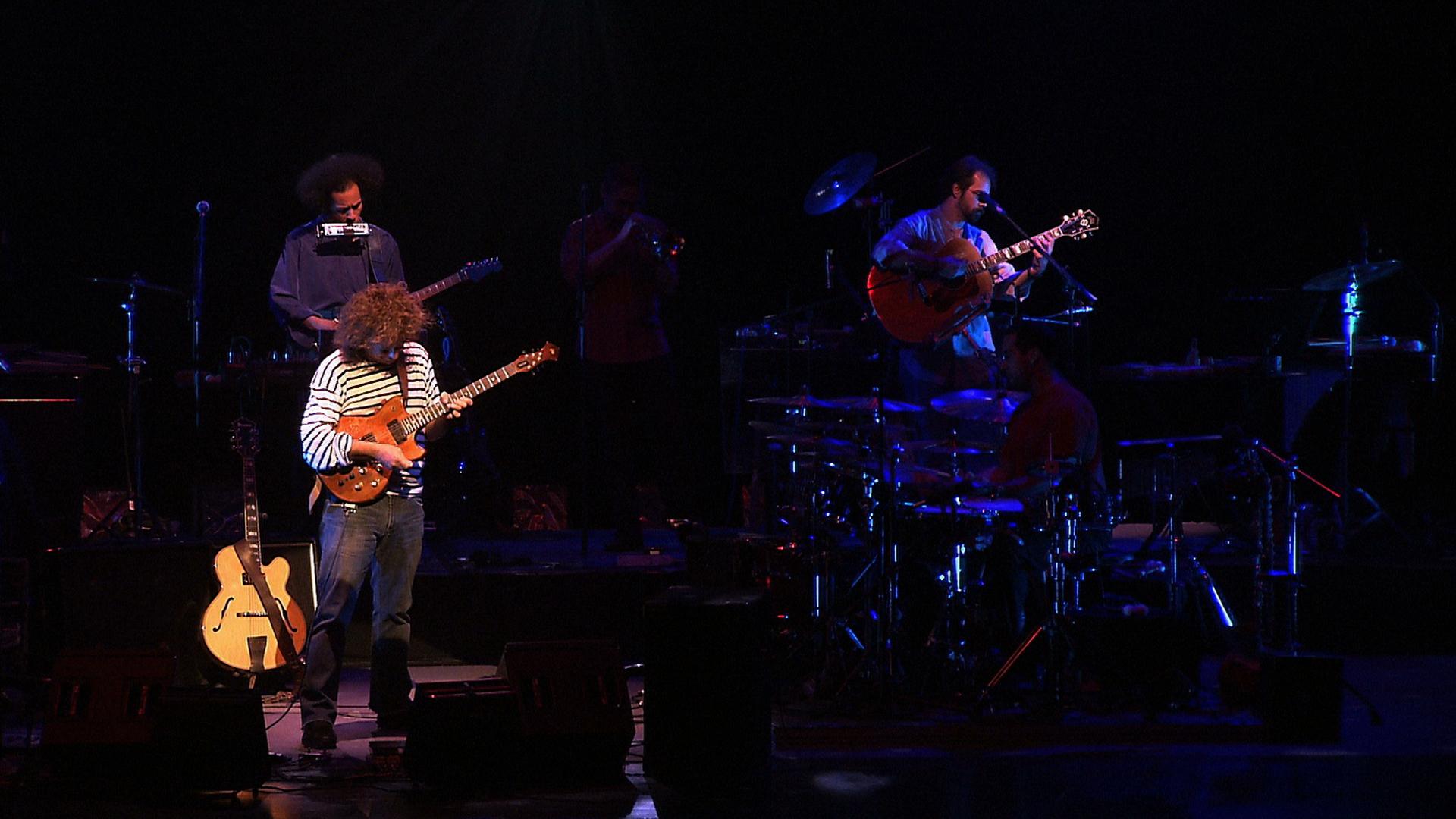 Pat Metheny performs on a dimly lit stage, featuring a guitarist at the front, a drummer, a bassist, and a trumpeter in the background.