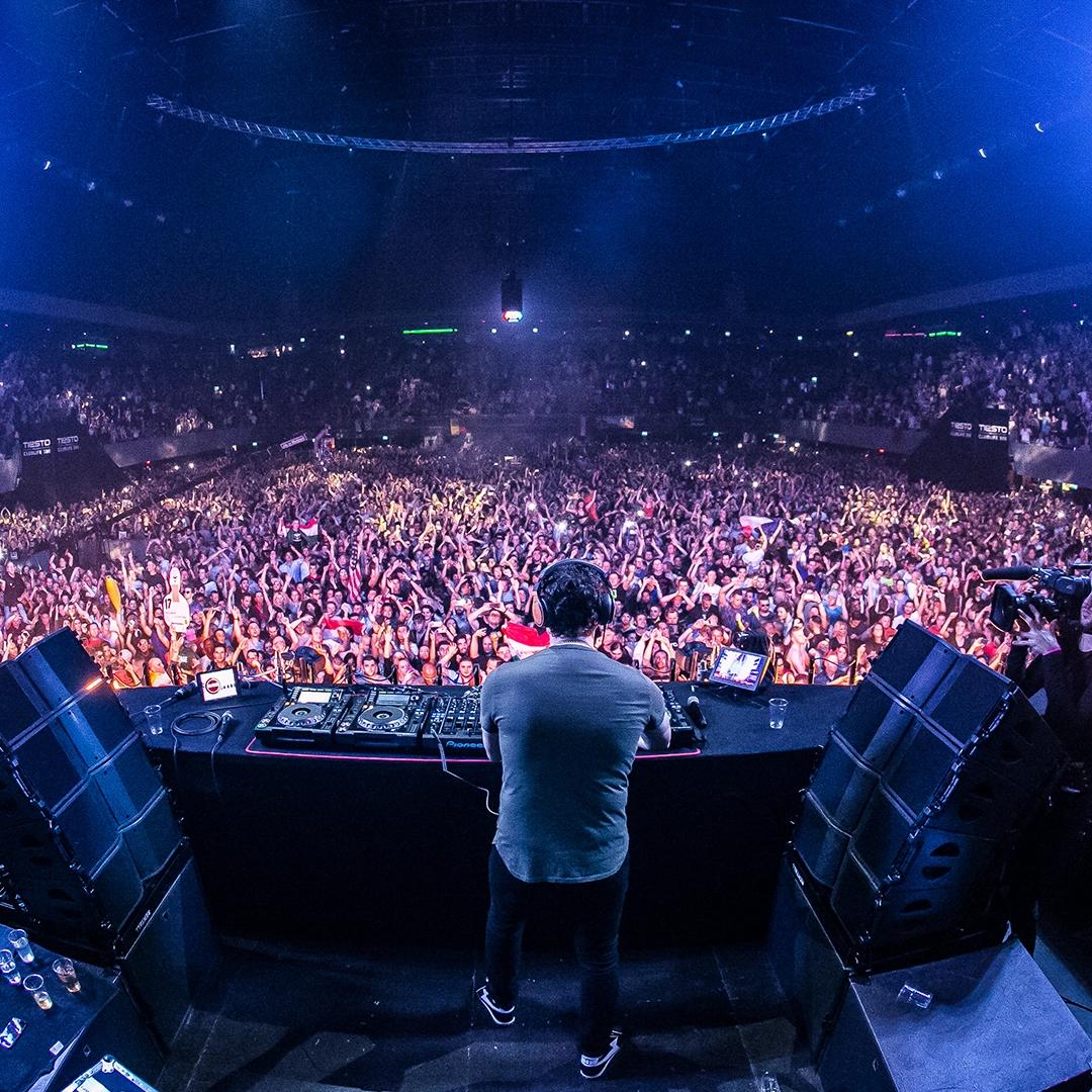 Tiësto performs on stage at a packed club or concert venue, with a cheering crowd in front and bright blue lights illuminating the scene.