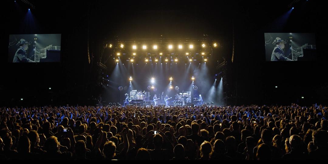 A large crowd at a Toto concert, illuminated by stage lights, watches a band perform on stage. Two large screens on either side display a close-up of one of the performers.