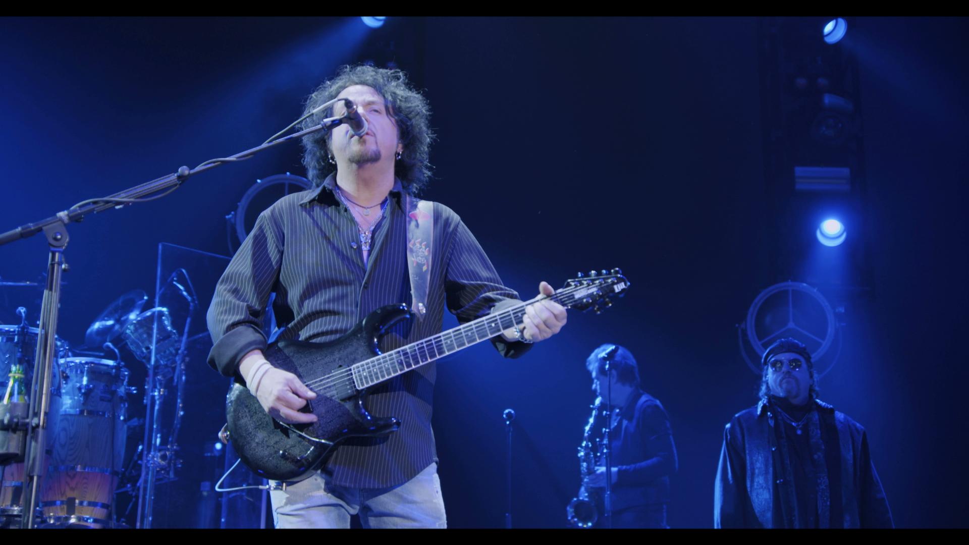 Member of Toto playing an electric guitar and singing into a microphone on a dark stage, accompanied by other musicians in the background.