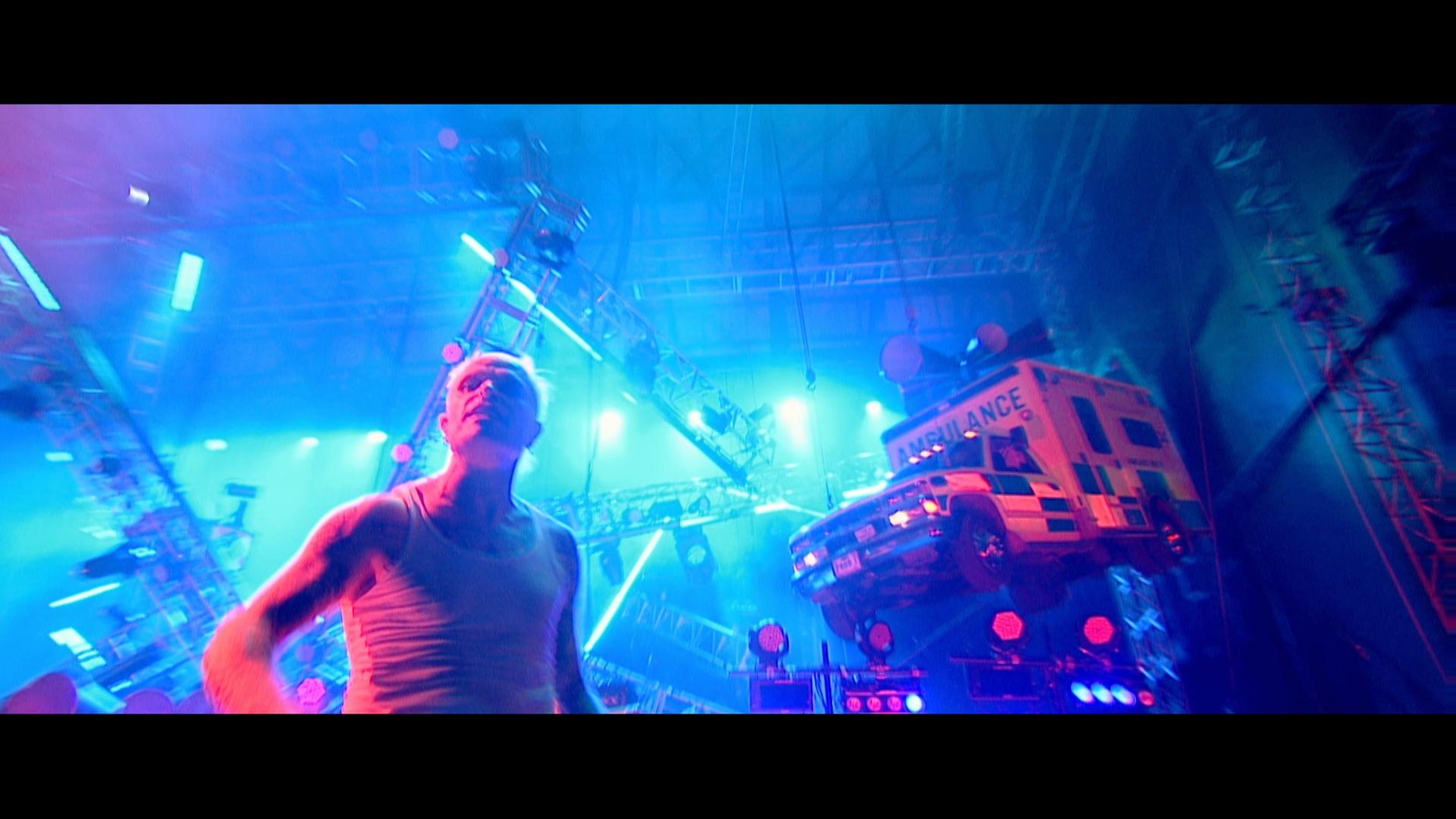 Kieth Flynn from the prodigy dressed in a white tank top stands on a stage with bright blue and purple lighting. An ambulance is suspended in the air in the background as part of the stage setup.