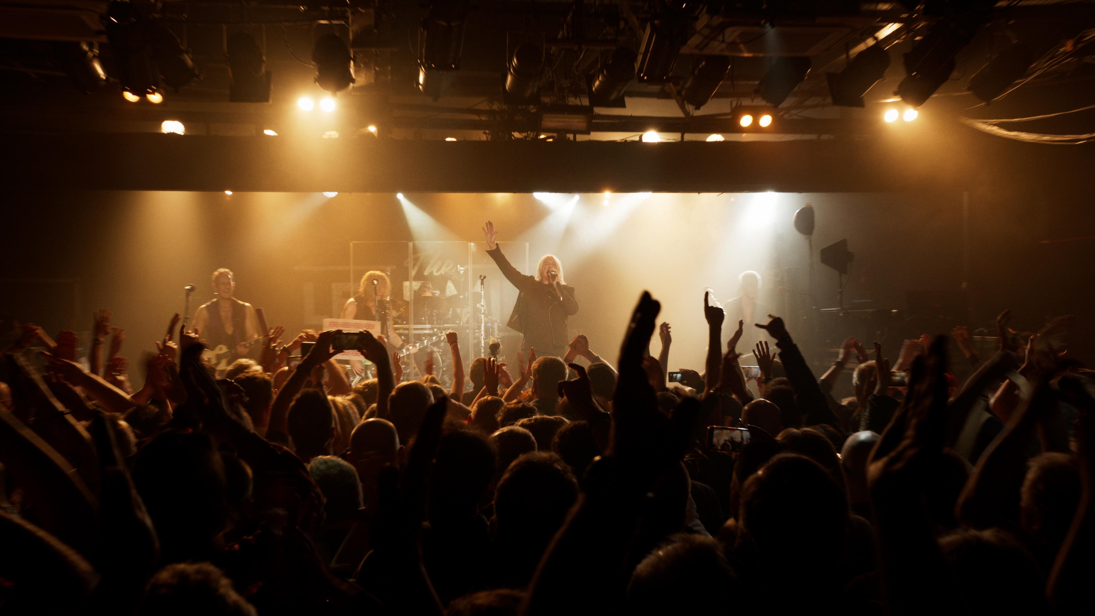 Def Leppard performs on stage with bright lights, facing a large, cheering crowd.