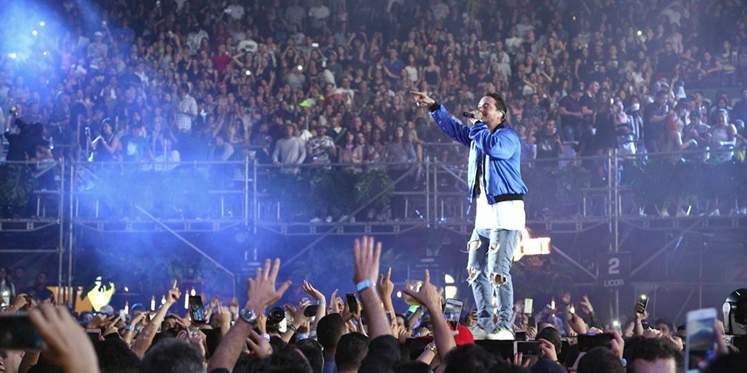 J Balvin in a purple jacket sings on stage to a large crowd at an outdoor concert, surrounded by raised hands and lit cell phones against a backdrop of stands filled with spectators.