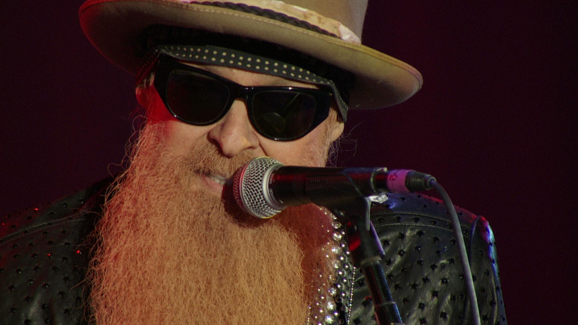 Billy F Gibbons with a long beard, wearing sunglasses, a hat, and a bandana, sings into a microphone on stage.