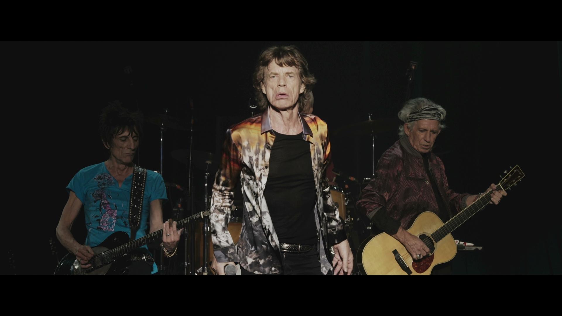 The Rolling Stones perform on stage, with one singing into a microphone and two others playing guitars. The background features a drum set.