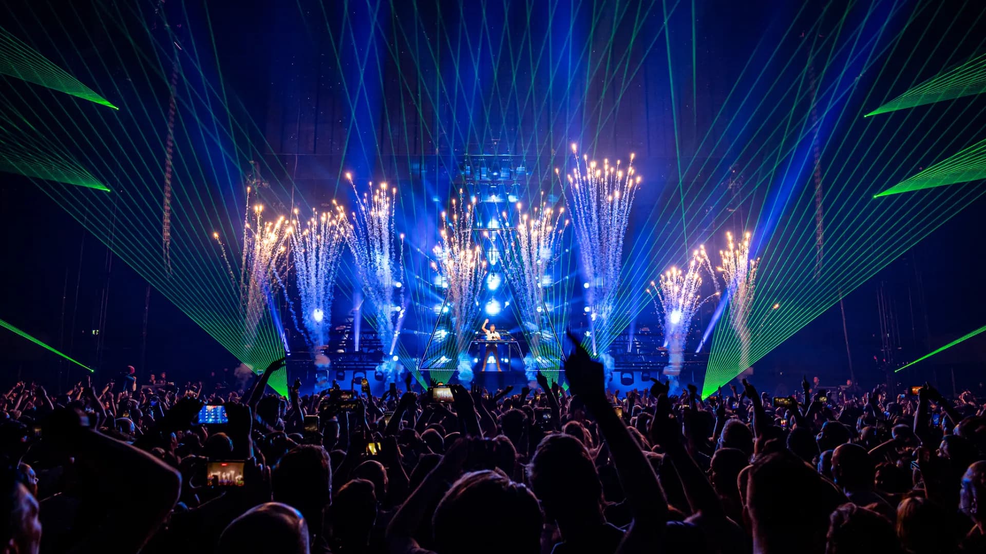 Concert audience enjoying a live performance at Ziggo Dome with bright stage lights, green laser beams, and pyrotechnic effects in the background.