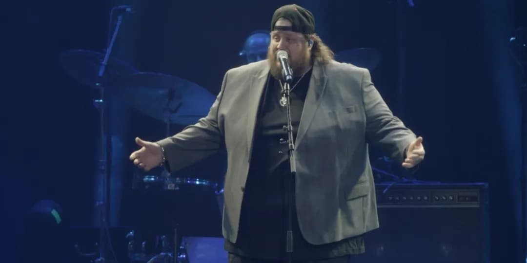 A person wearing a gray blazer and black cap sings or speaks into a microphone on a dimly lit stage, with drums and other instruments visible in the background.