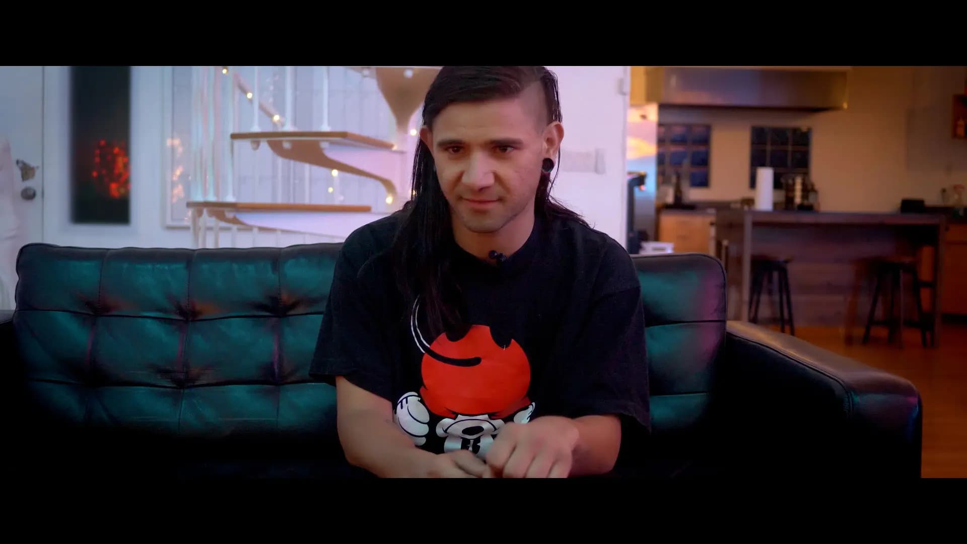 Bass Impact contributor with long hair and earrings sitting on a black leather couch, wearing a black shirt with a red graphic, in a room with modern decor and string lights.