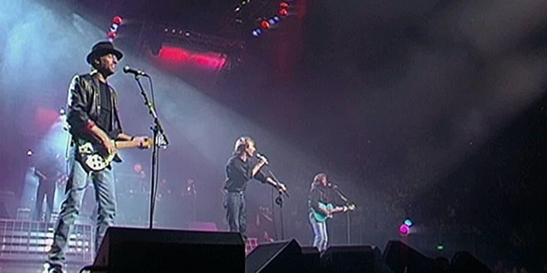 Three musicians perform on a stage with microphones and guitars under colorful stage lights, with a smoky atmosphere and audience in the background.