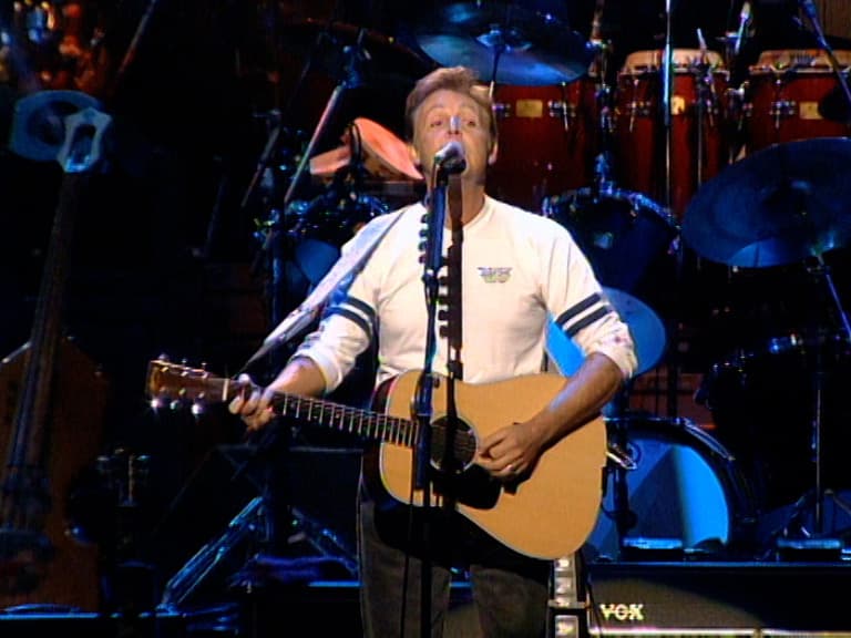 Paul McCartney stands on stage playing an acoustic guitar and singing into a microphone, with drums and stage lights in the background.