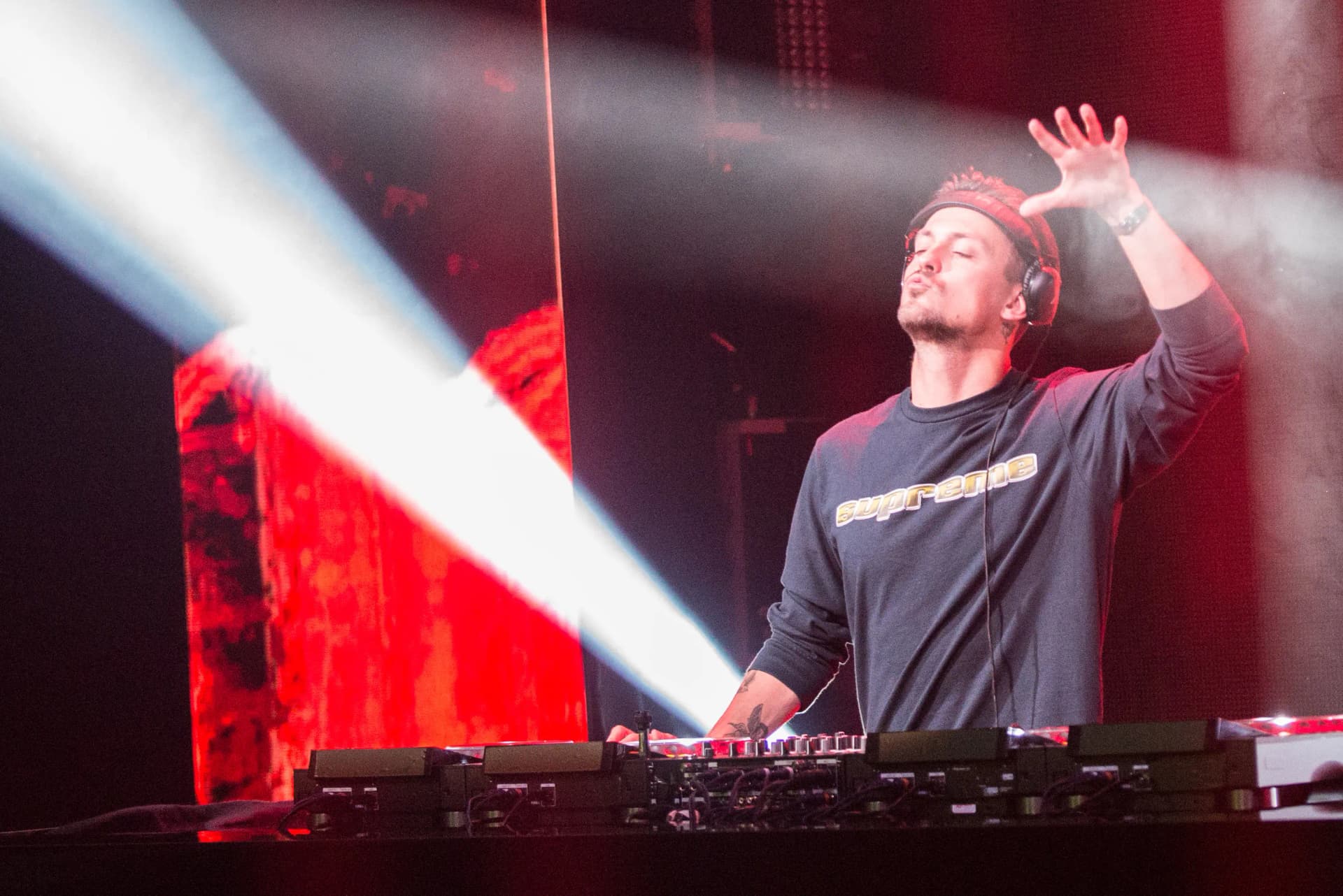 Julian Jordan wearing headphones, standing at a mixing console, illuminated by colorful stage lights, with one hand raised.