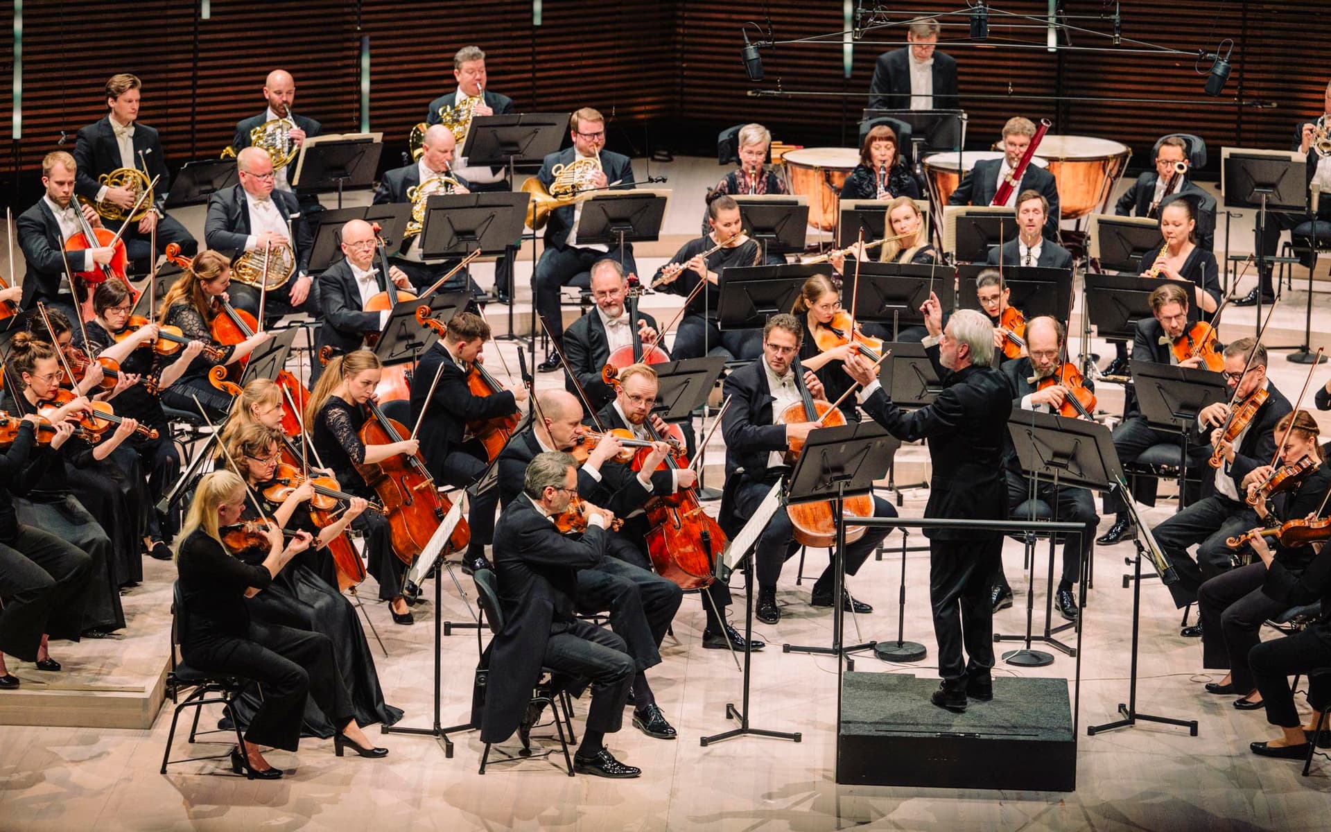 An orchestra performs on stage, with musicians playing various string, woodwind, brass, and percussion instruments under the direction of a conductor.